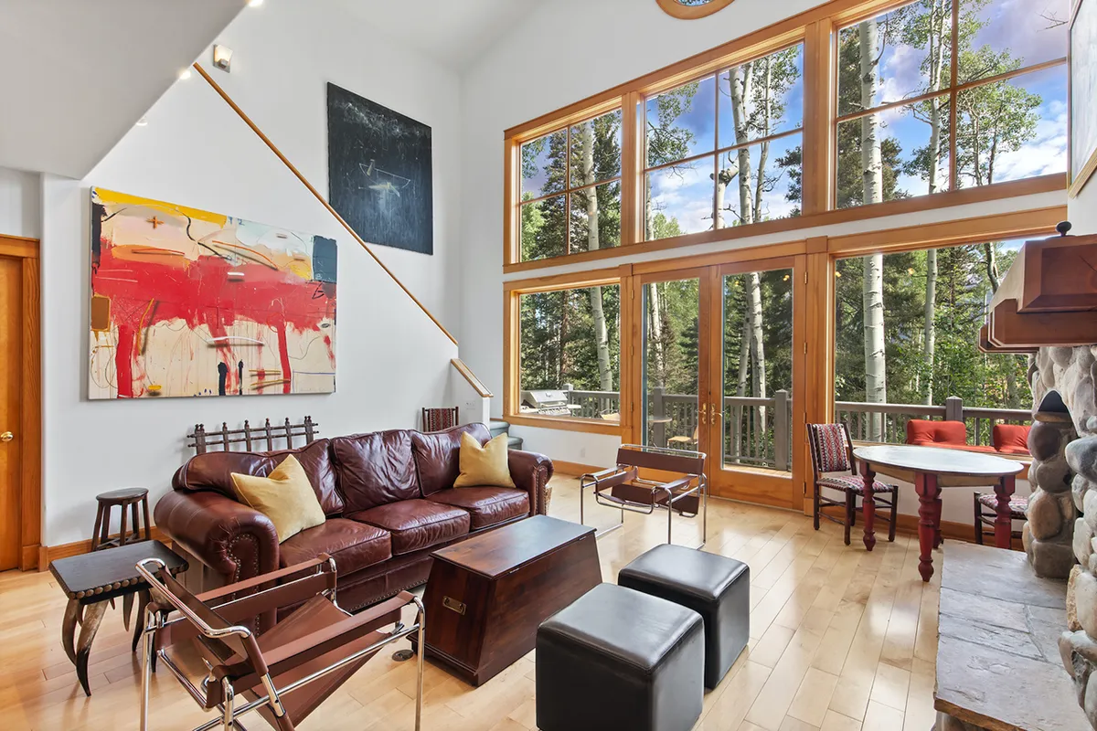 A spacious, light-filled living area overlooks a brown leather sofa and wooden coffee table, with expansive windows revealing lush forest views. A colorful abstract painting adds a creative, open feel, suggesting the inspiration and energy that flow throughout the space below the loft.