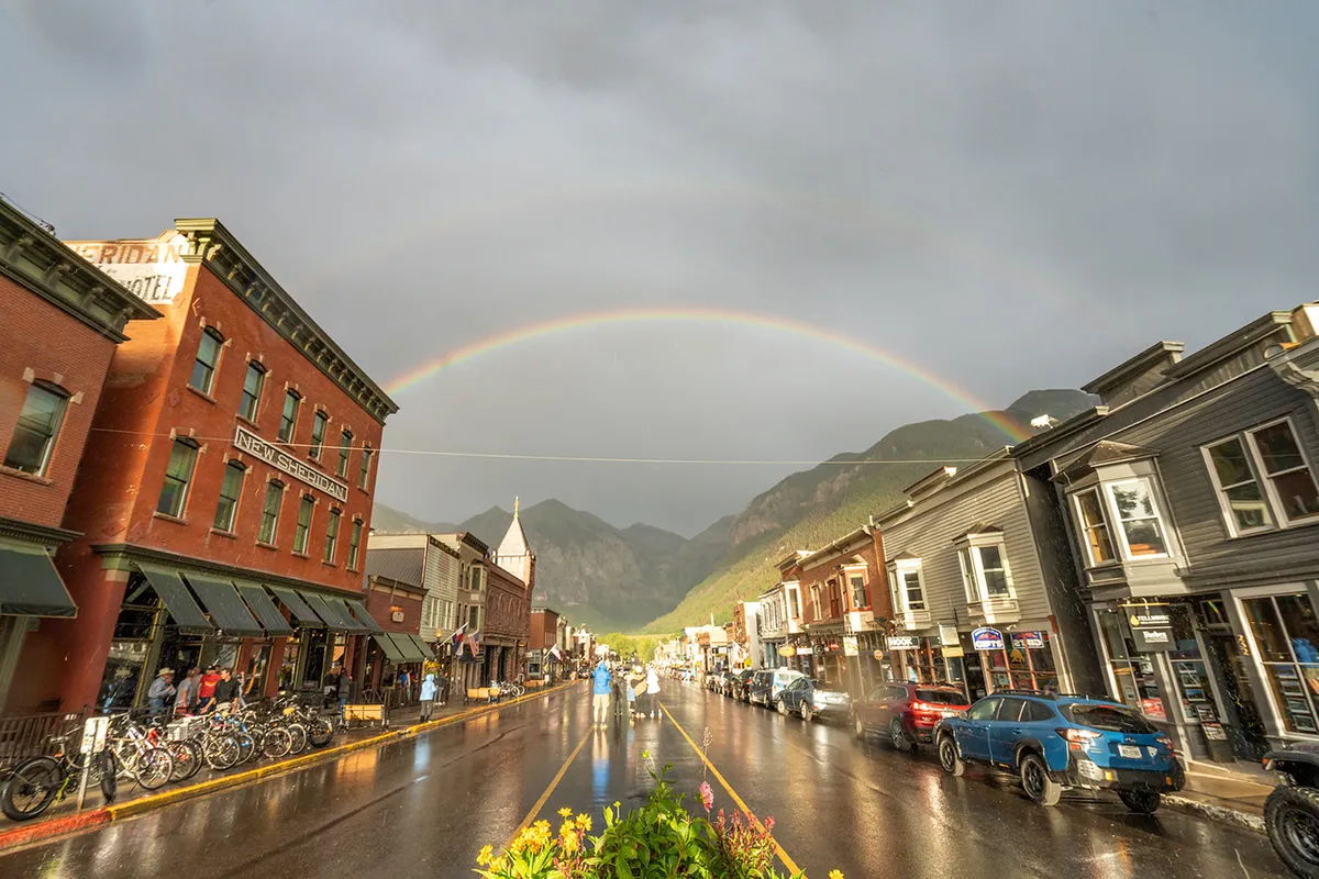 A charming mountain town street scene with historic buildings lining a wet road, a vibrant rainbow arching overhead, and majestic mountains in the background. The scene captures couples strolling, shopping, or pausing to admire the architecture and rainbow after a gentle rain.