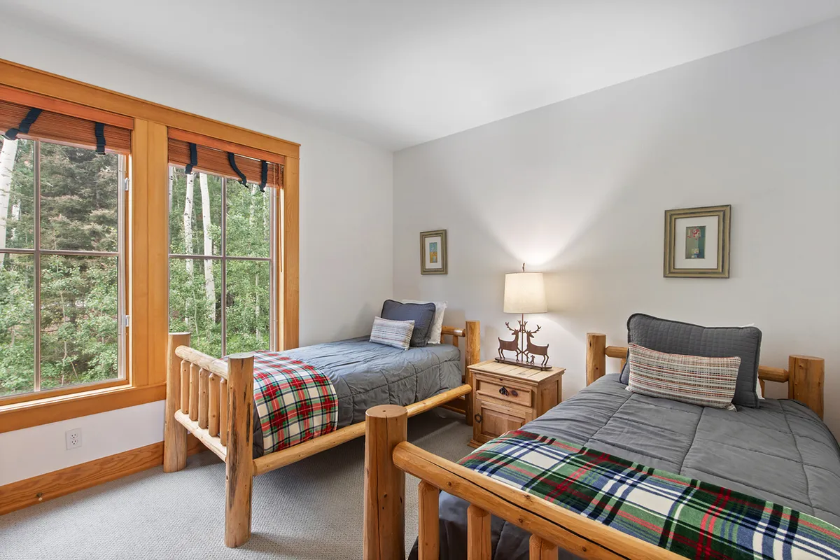 A bright bedroom with two wooden beds, large windows letting in natural light, and a bedside lamp. The space feels warm and inviting—perfect for children, siblings, or guests to share.