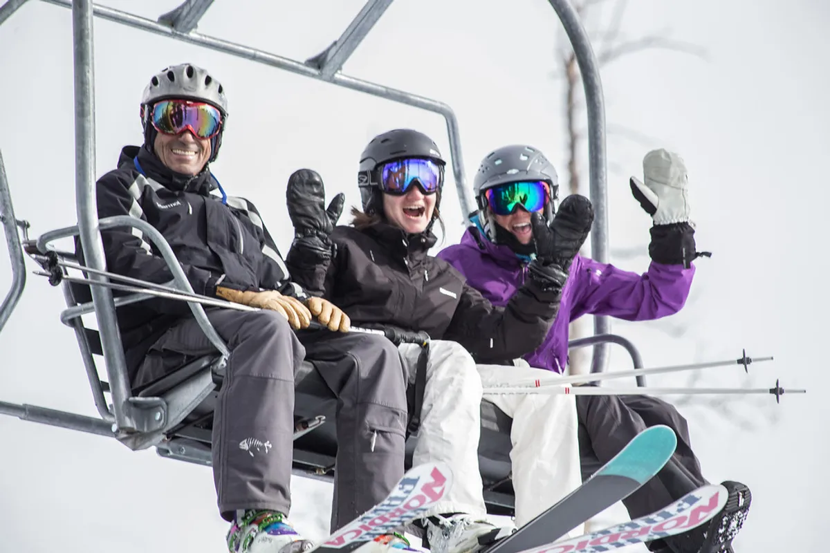 Three skiers ride a chairlift above snowy terrain, with ski gear visible and a cheerful, sociable mood. The snow-covered landscape hints at the nearby ski runs, underlining the chalet’s proximity to the slopes and the vibrant après-ski culture.
