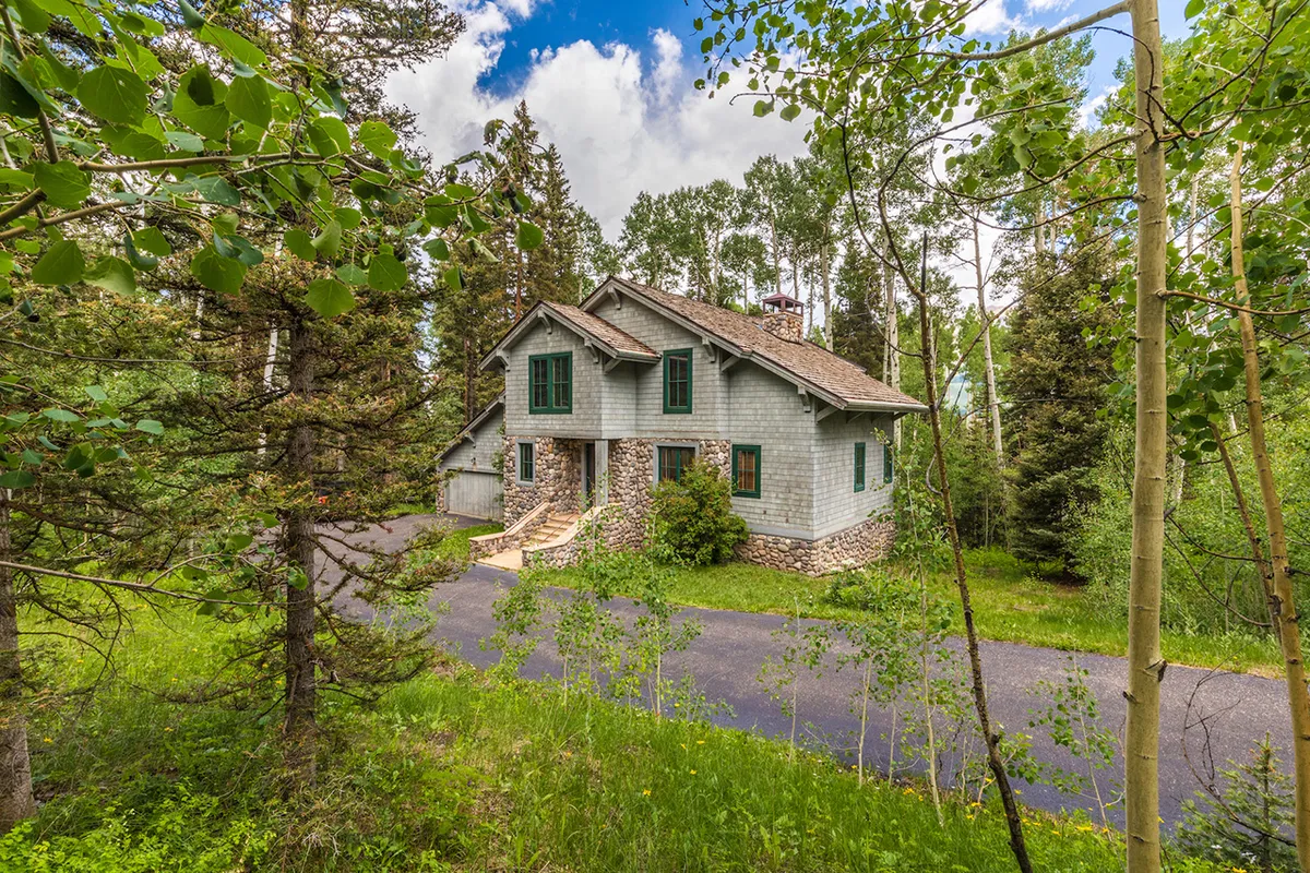 The exterior of a cozy house surrounded by tall trees, with a stone pathway leading up to the entrance. The setting is peaceful and inviting, hinting at outdoor adventures and relaxation in nature.