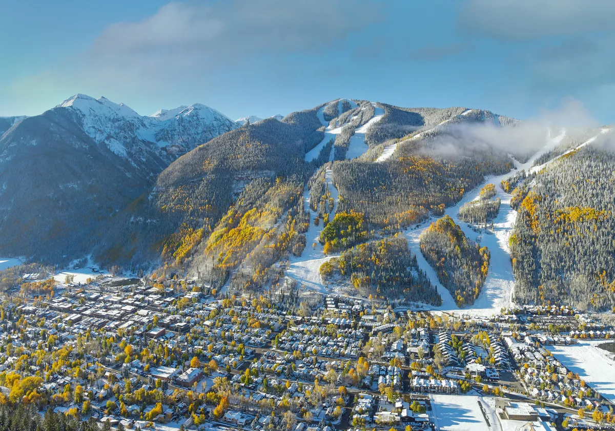 A panoramic view of snow-capped mountains, ski slopes, colorful autumn trees, and the quaint town below. The landscape captures both the adventure of the slopes and the tranquility awaiting at home or in town.