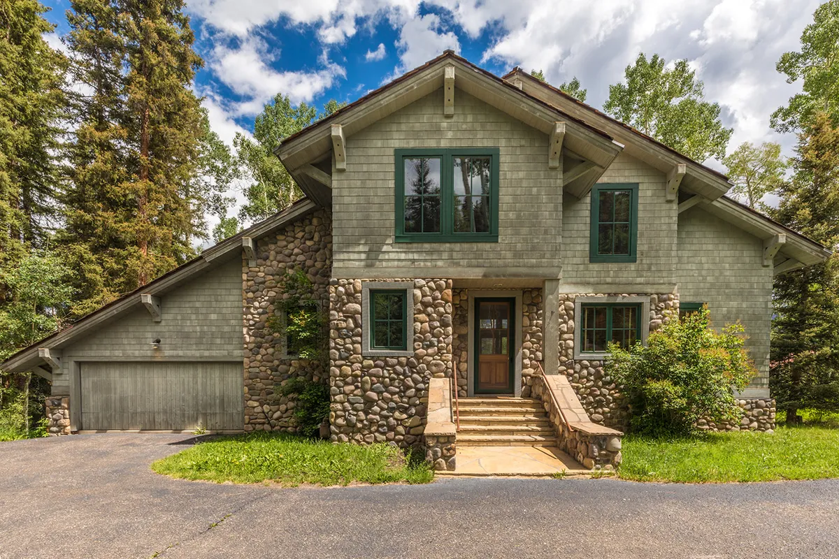 The front entrance of the chalet is framed by a beautiful stone facade and large windows, all surrounded by lush alpine trees. This image emphasizes the welcoming aspect of the property and hints at both comfort and convenience, as the garage and easy outdoor access are implicit in the visual.