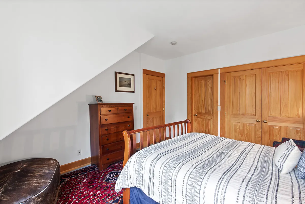 A warmly lit bedroom featuring a comfortable bed with plush bedding, a chest of drawers, a decorative rug, and colorful paintings on the walls. Soft sunlight filters in, creating a relaxed and cozy environment ideal for restful nights or peaceful mornings.