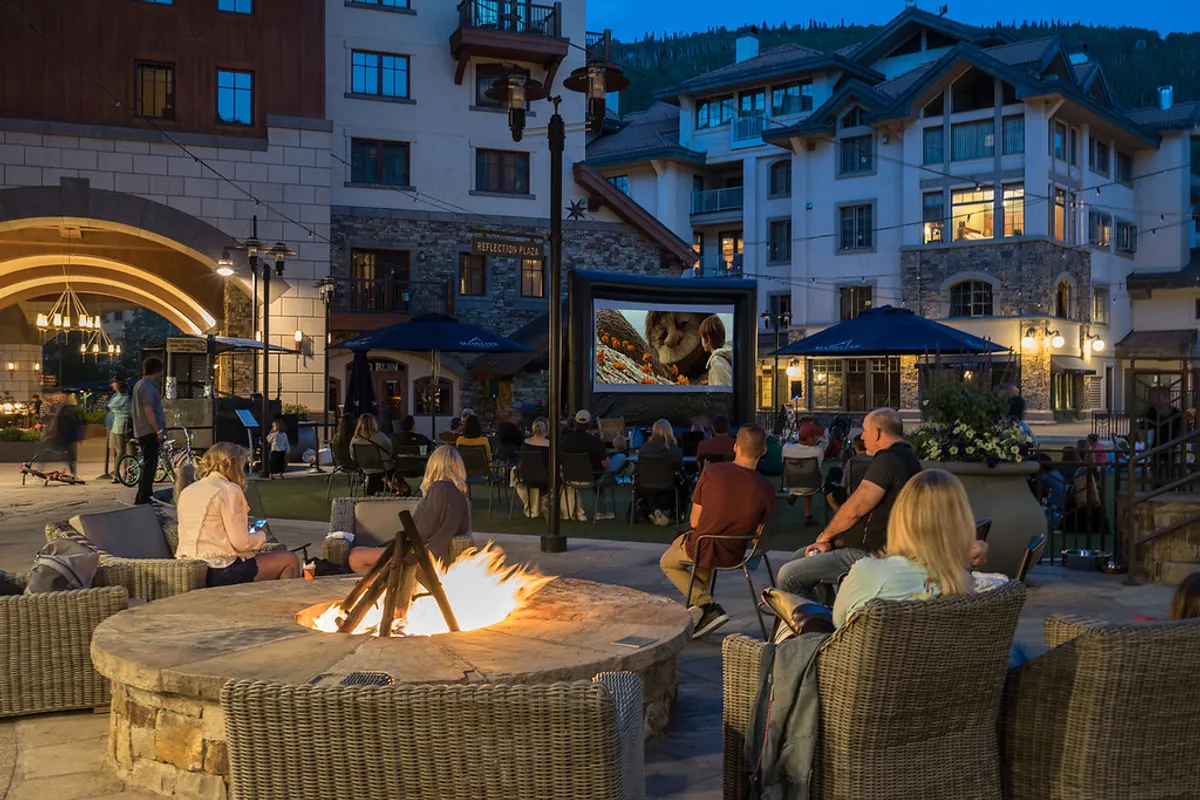 An outdoor communal area featuring a fire pit, comfortable seating, and an outdoor movie screen, surrounded by alpine architecture. The setting is primed for group gatherings, storytelling, and relaxing under the evening sky.