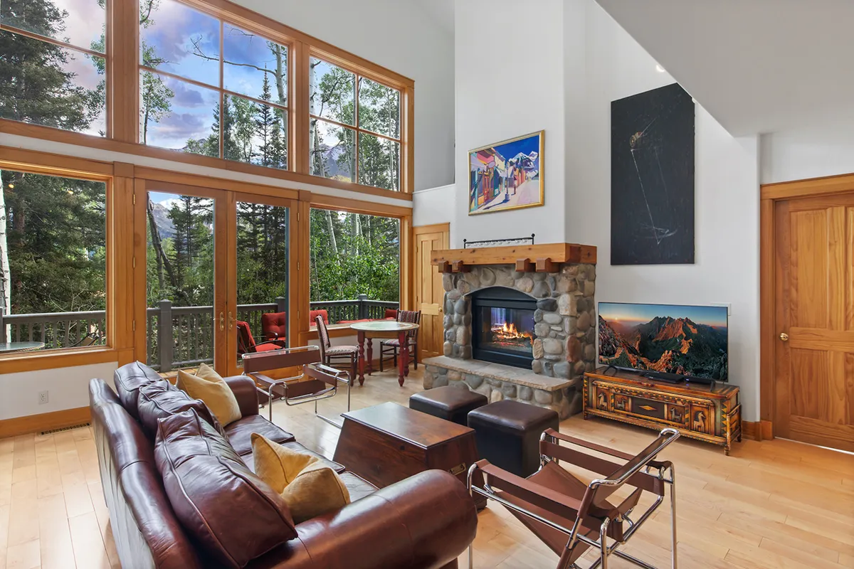 The image showcases a warm, inviting living space featuring a crackling fireplace, plush leather sofa, and large windows framing stunning mountain views. The setting is perfect for families or friends to gather, relax, and enjoy serene winter evenings together.