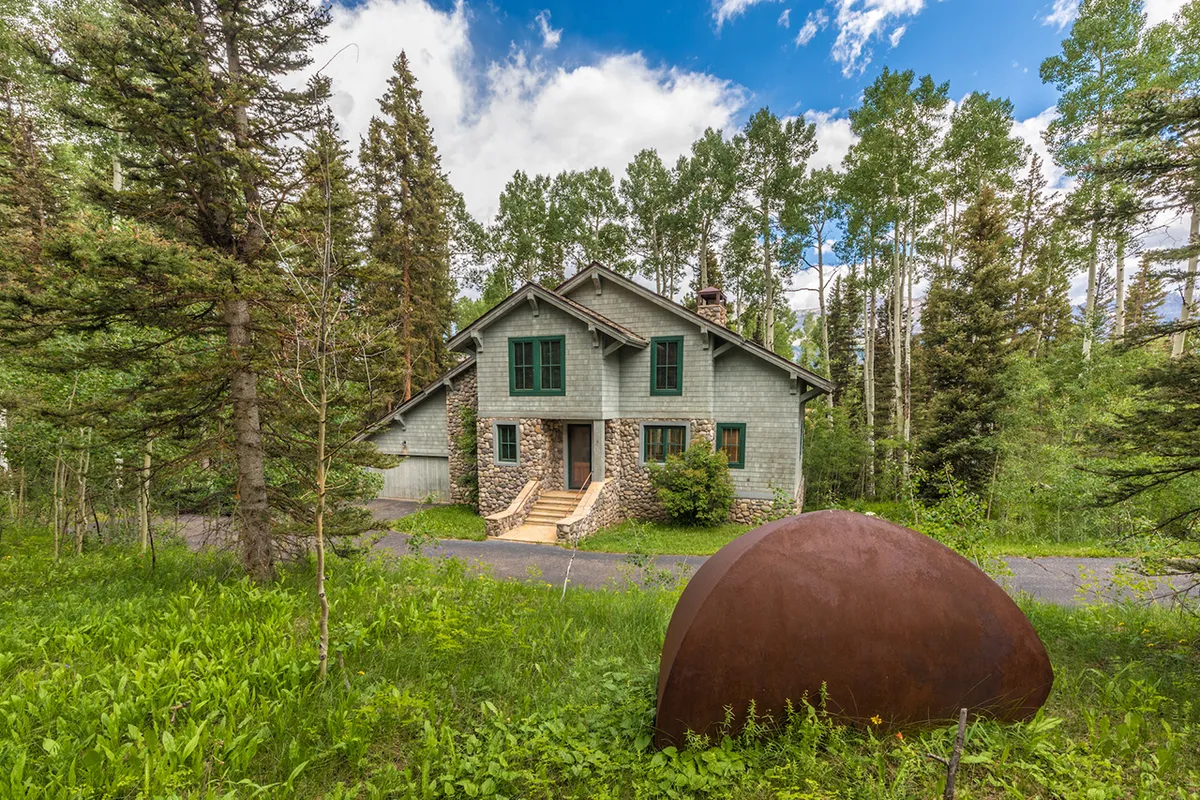 The house sits surrounded by lush trees and greenery, with a rustic metal sculpture adding character to the peaceful exterior. The environment feels calm and inviting, ideal for family gatherings and outdoor enjoyment.
