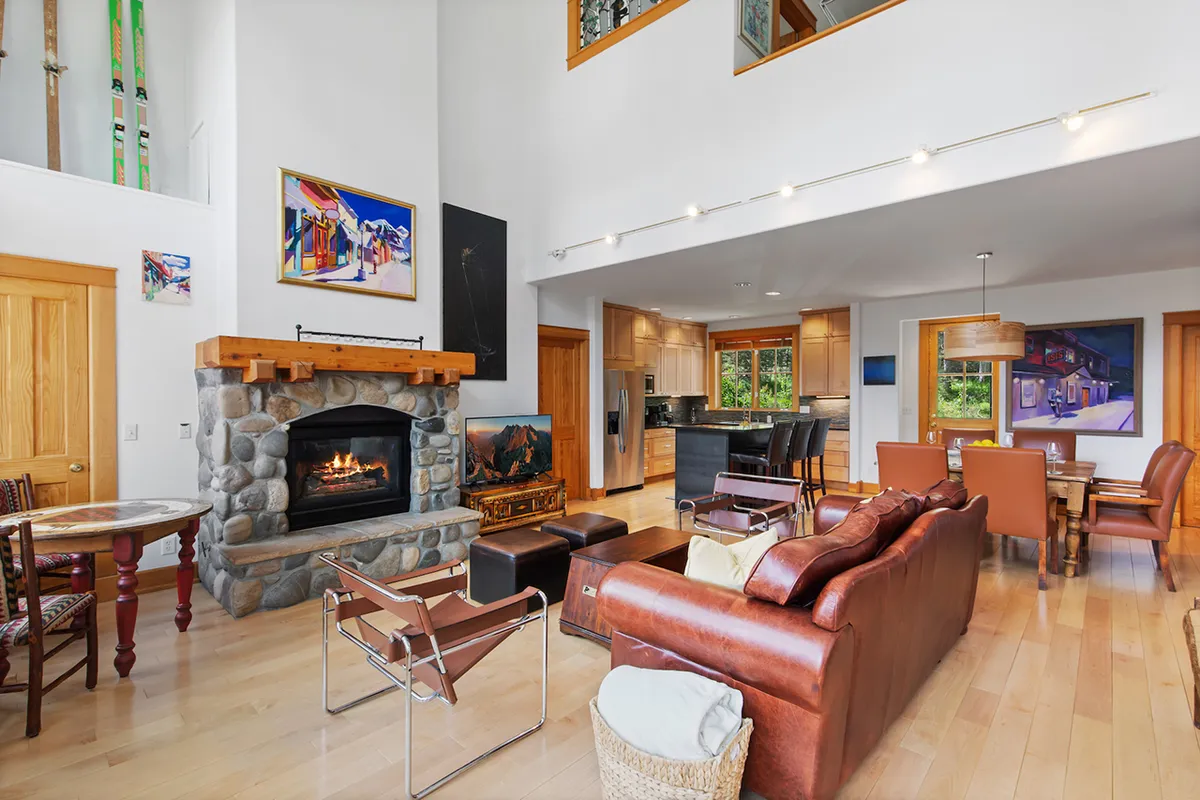 This image features a welcoming living area with a stone fireplace, large windows, a leather sofa, and a wooden dining area. Sunlight streams through the expansive windows, illuminating the cozy and spacious setting. The space radiates warmth and comfort, inviting gatherings around the fireplace with beautiful views beyond.