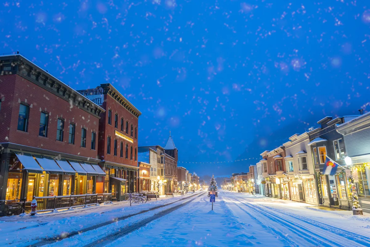A snow-dusted village street with warmly decorated storefronts and a glowing Christmas tree in the distance, evoking the joy of winter strolls, holiday shopping, and cozy café visits.