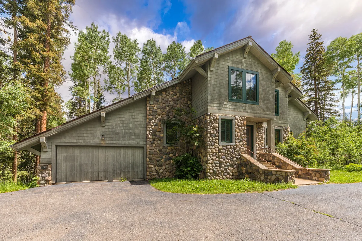 A welcoming chalet-style exterior with a stone facade, large windows, and a front porch set amid towering trees. The inviting steps and warm lighting hint at the comfort inside, while the natural setting promises outdoor adventures and gatherings.