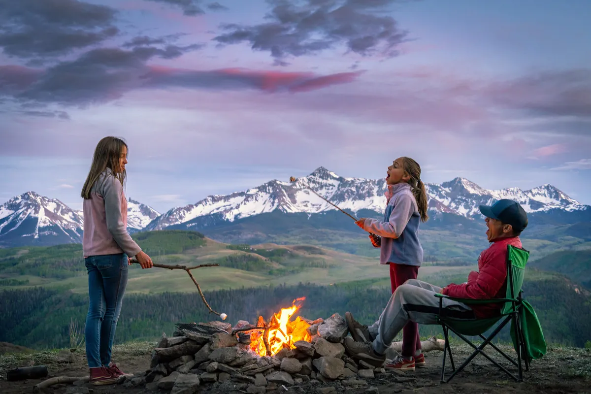 Children and adults gather around a campfire, roasting marshmallows with mountains in the background. The mood is joyful and communal, capturing the essence of family-friendly outdoor activities and making lasting memories together.