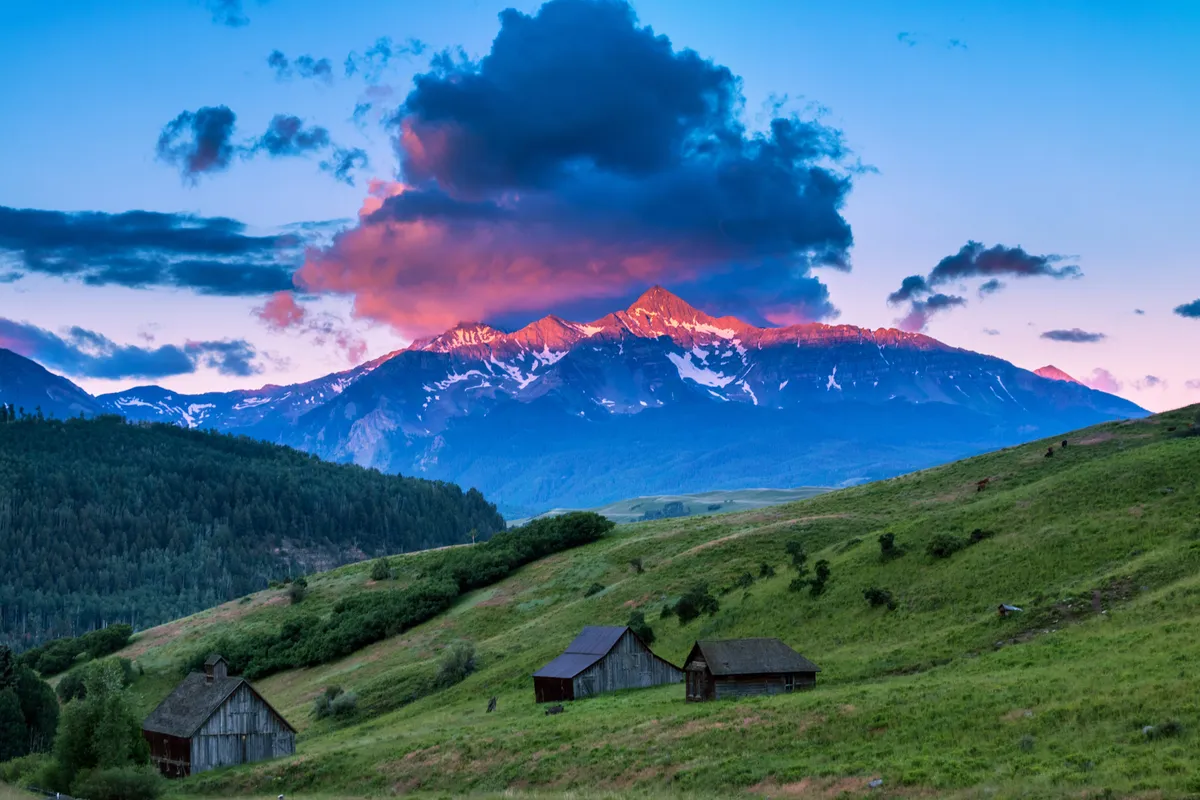 A sweeping view of majestic mountains under sunset-hued clouds, with rustic wooden cabins nestled among lush green hills in the foreground. The scene exudes tranquility, with expansive vistas inviting thoughts of fresh mountain air and endless outdoor possibilities.