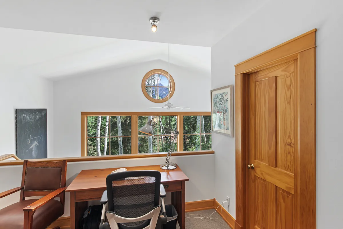 A focused view of a wooden desk positioned beneath a distinctive round window, paired with a comfortable chair and illuminated by a desk lamp. The serene backdrop visible through the window hints at treetop or potential mountain views, setting a tranquil tone perfect for work or study.