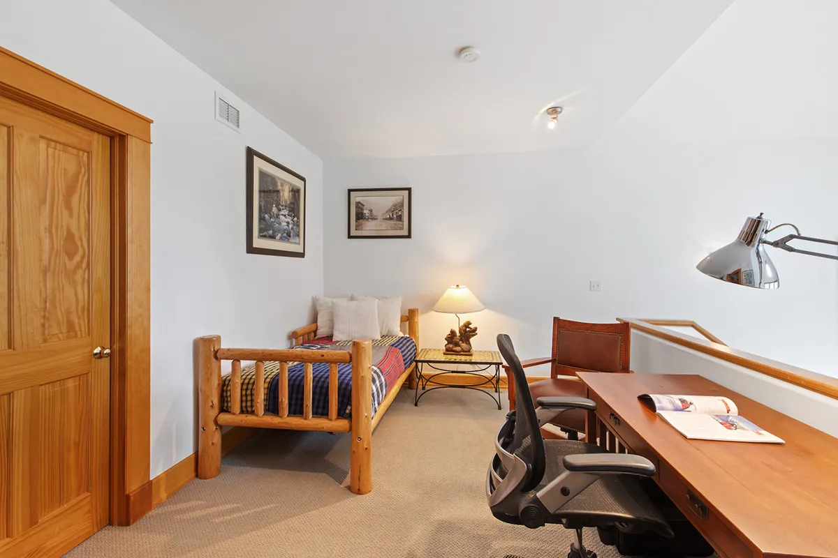 A cozy nook features a twin bed with colorful bedding alongside a functional desk with an ergonomic chair and a warm table lamp. The setup radiates comfort and versatility, highlighting the ability for both relaxation and productivity within a single area.