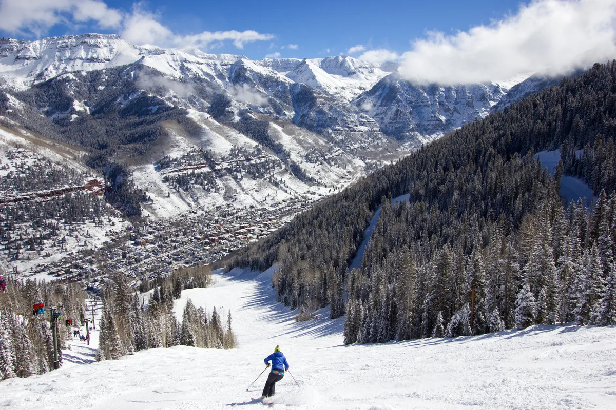 A sweeping view of snow-covered mountains with a skier descending the slope, dense pine forests, and a quaint town nestled in the valley below. The image captures the excitement of skiing with breathtaking panoramic scenery.