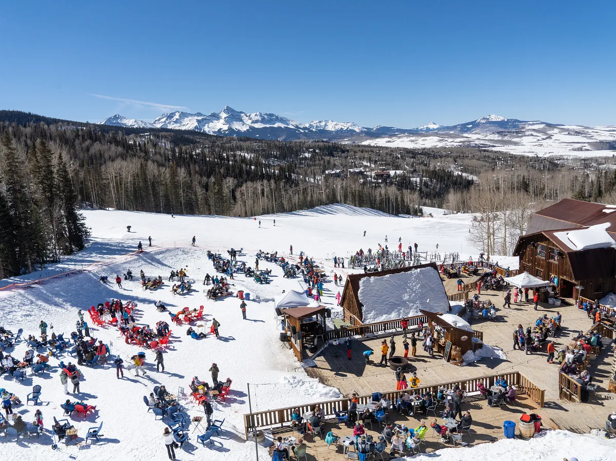 A lively scene featuring people skiing down snowy slopes, a mountain range in the background, an outdoor seating area, and a welcoming lodge. The setting is energetic, social, and festive, with the sun casting warm light over the landscape.