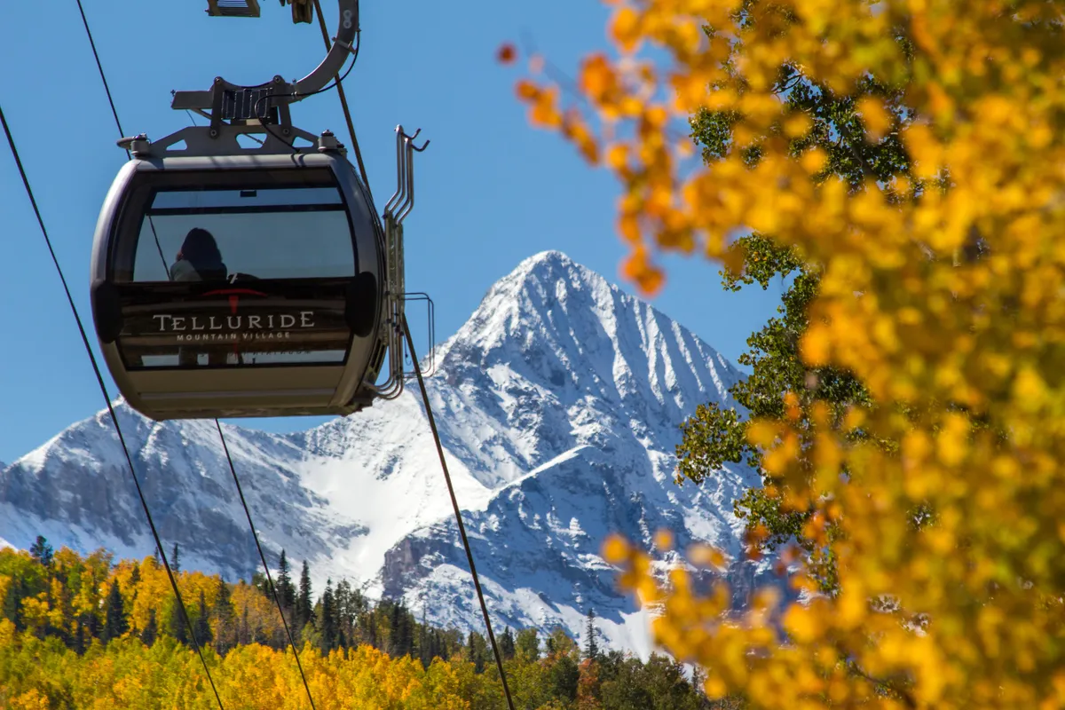 A gondola lift glides over a mountain landscape, with autumn foliage coloring the valleys and a snow-capped peak in the background. The scene is vibrant and scenic, evoking the sense of adventure and easy access between Mountain Village and Telluride.
