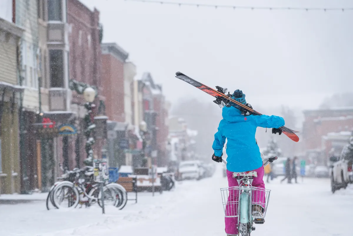 A lively snow-covered street scene featuring a person on a bike carrying skis, set against historic buildings. The setting is energetic and cheerful, suggesting the proximity to both winter sports and vibrant village life.